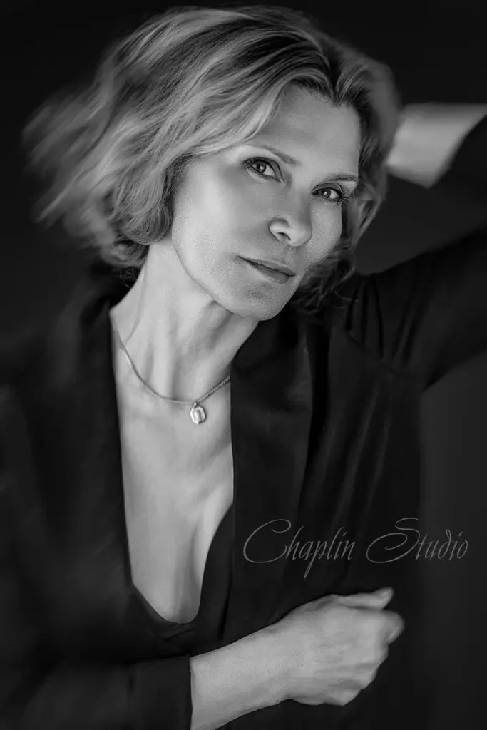 A striking black and white portrait of a woman with short, light hair, wearing a dark, open-neck top and a necklace, looking directly at the camera with a thoughtful expression, captured by Chaplin Studio.