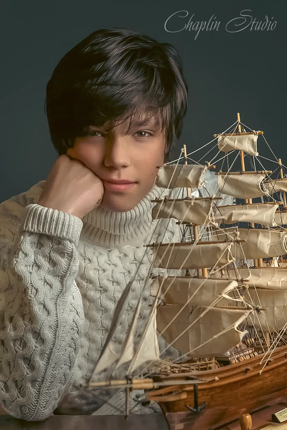 A child with dark hair, wearing a cream sweater, poses thoughtfully next to a detailed model ship, captured in a creative portrait by Chaplin Studio in Yorkshire.