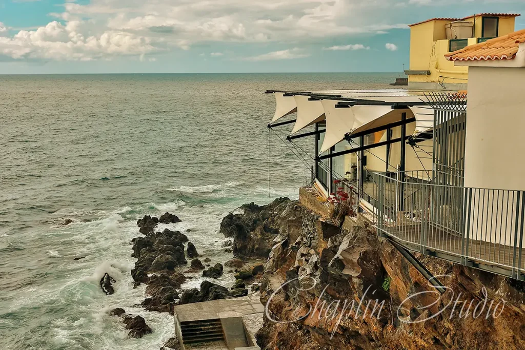 A modern coastal restaurant built into a cliffside overlooking a rough ocean with waves crashing on rocks, featuring white awnings and a balcony, captured by Chaplin Studio.