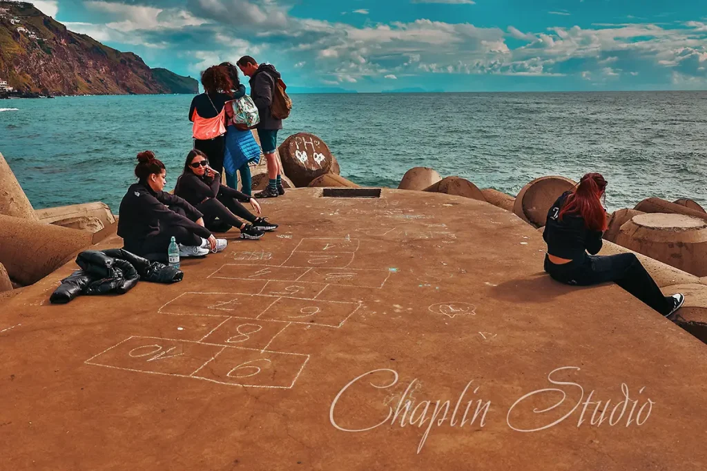 A group of people relaxing and interacting on a concrete pier by the ocean, with a hopscotch game drawn on the ground, and coastal mountains in the background under a cloudy sky, captured by Chaplin Studio.