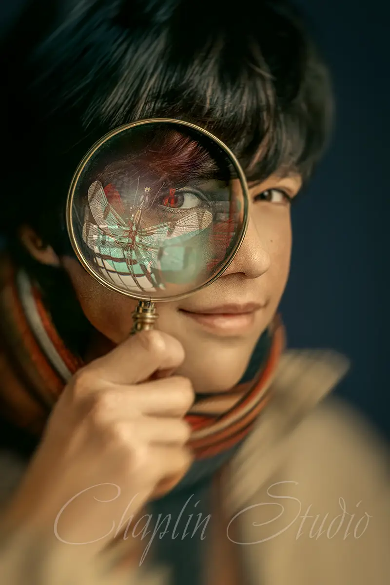 A creative portrait of a child holding a magnifying glass, with a detailed reflection of a dragonfly in the lens, captured by Chaplin Studio. This image highlights curiosity and artistic vision.