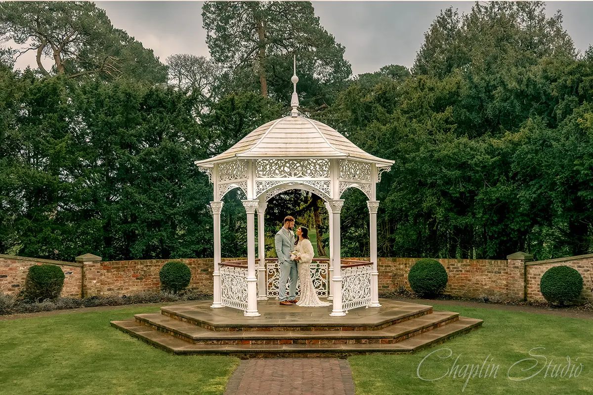 An elegant wedding couple standing under a white ornate gazebo in a lush green garden, surrounded by trees and a brick wall, captured by Chaplin Studio, a Yorkshire Wedding Photographer.