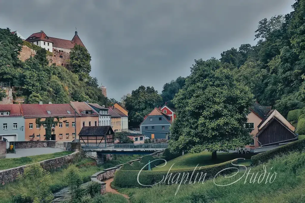 A picturesque European village nestled in a valley with colorful houses and a historic castle on a lush green hilltop, featuring a bridge and winding path, captured by Chaplin Studio.