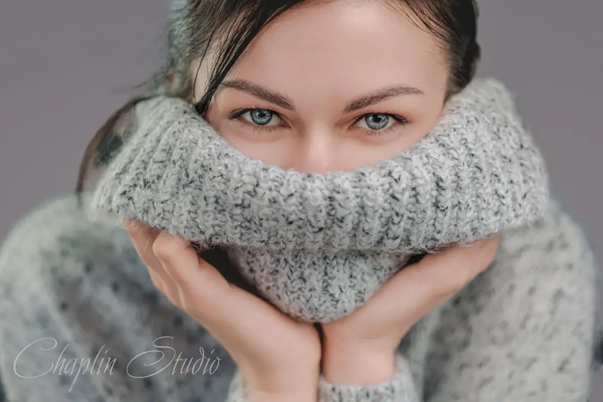 A close-up portrait of a woman with striking blue eyes, partially obscured by a cozy grey knitted turtleneck sweater, conveying a sense of warmth and mystery, captured by Chaplin Studio.