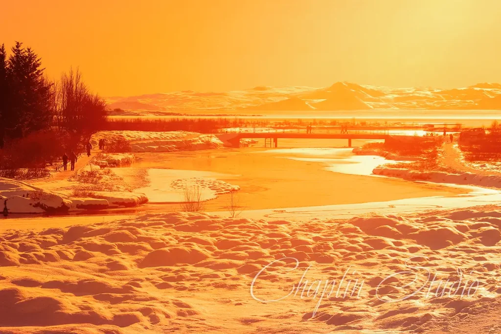 A stunning winter landscape bathed in golden hour light, showing a snow-covered foreground, a partially frozen river, a bridge with distant figures, and mountains under a bright sky, captured by Chaplin Studio.