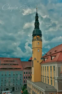 A prominent historic church tower with a black spire and yellow facade, rising above red-tiled roofs and traditional European buildings under a cloudy sky, captured by Chaplin Studio.
