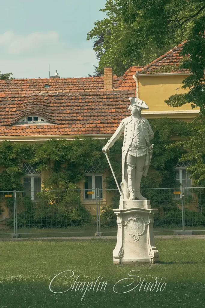 A white marble statue of a historical figure in a park, with a traditional European building with red-tiled roofs and green foliage in the background, captured by Chaplin Studio.