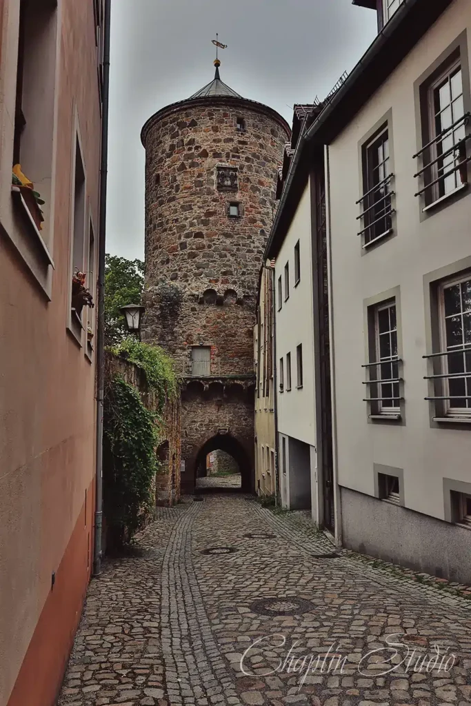 A narrow, historic cobblestone street leading to a tall, round stone tower with an arched entrance, flanked by old European buildings, captured by Chaplin Studio.