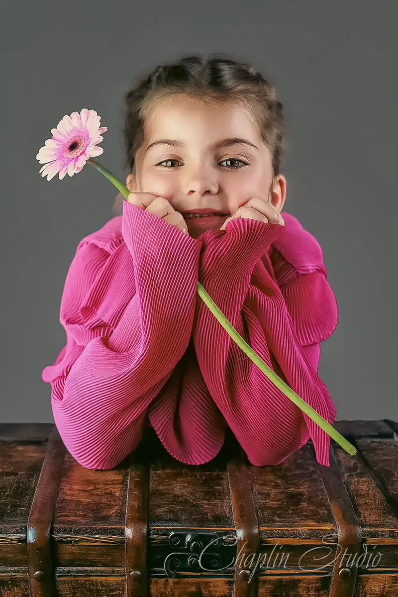 Little girl in a pink dress with a flower, leaning on an antique chest – fine art portrait by Chaplin Studio