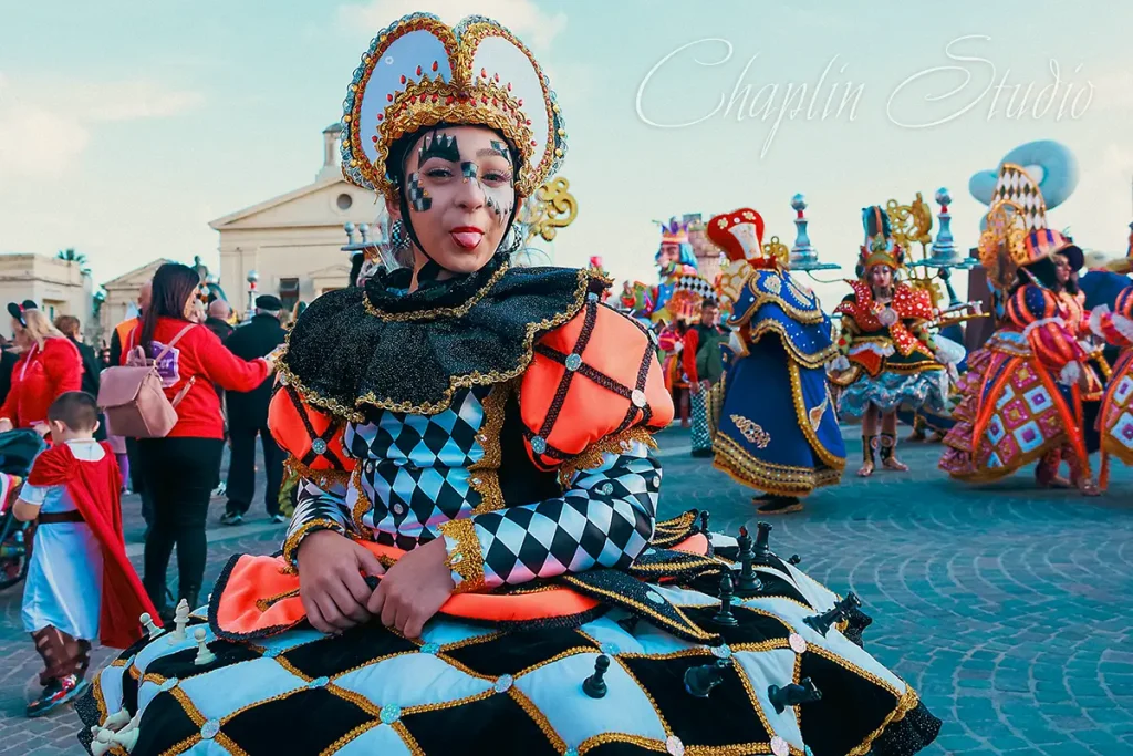 A person in an elaborate black, white, and orange chess-themed carnival costume with face paint, sticking out their tongue, surrounded by other costumed participants at a vibrant outdoor festival in Malta, captured by Chaplin Studio.