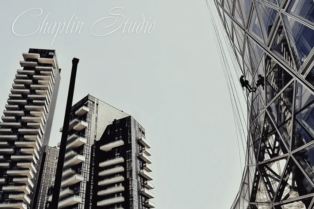 A low-angle view of modern skyscrapers with geometric glass facades, showing two window cleaners rappelling down the side of one building, under a bright sky, captured by Chaplin Studio.