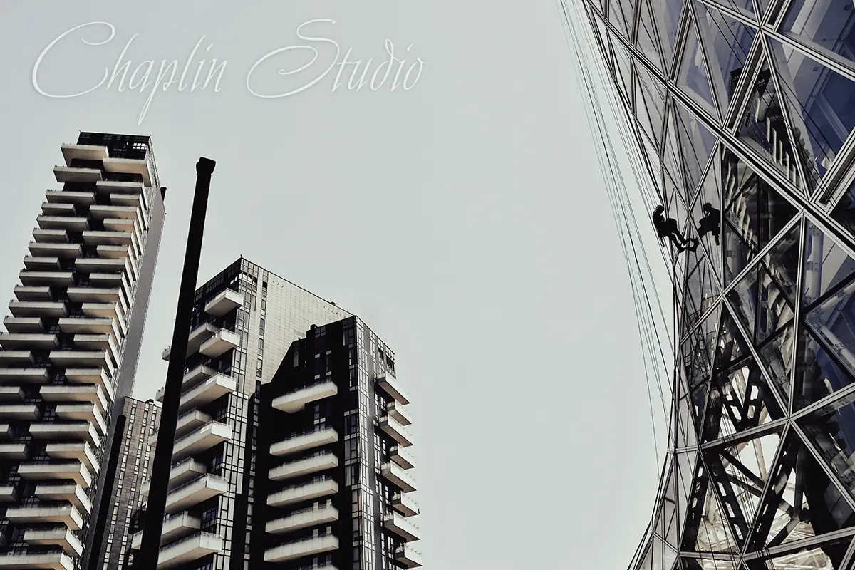 A low-angle view of modern skyscrapers with geometric glass facades, showing two window cleaners rappelling down the side of one building, under a bright sky, captured by Chaplin Studio.