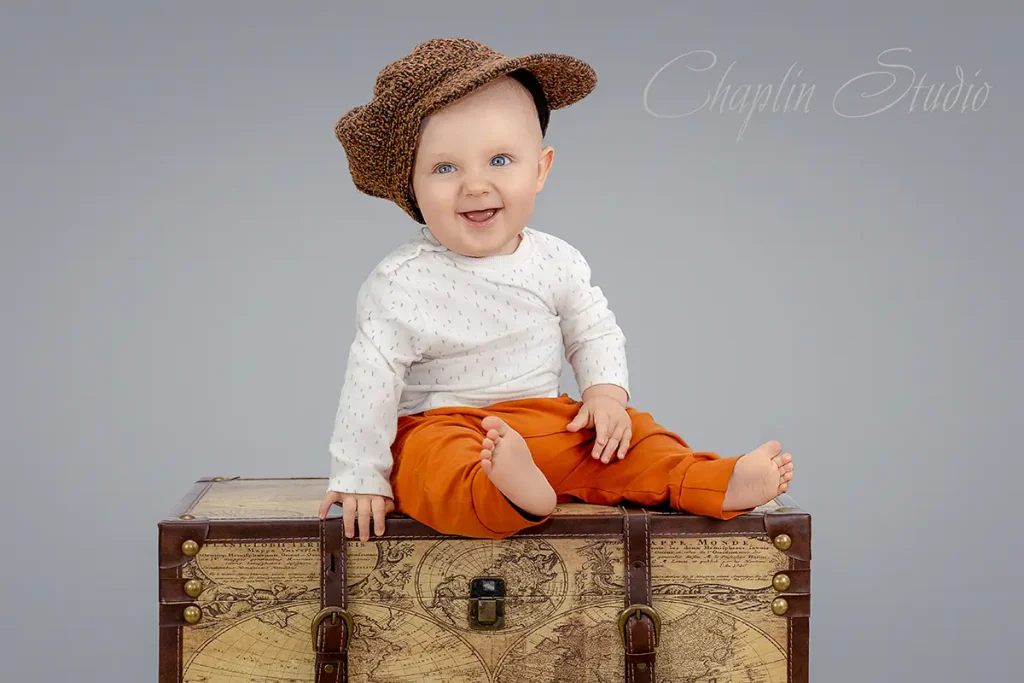 Smiling baby boy in orange trousers and knitted hat sitting on a vintage suitcase, photographed by Chaplin Studio Yorkshire.