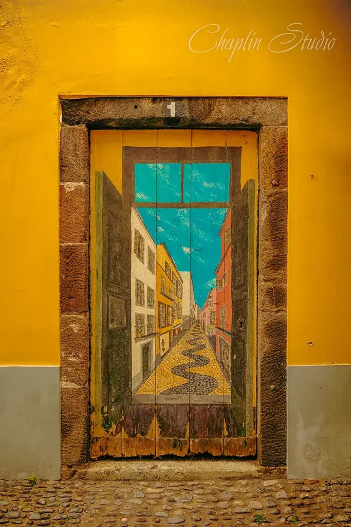 A vibrant yellow building wall with an old wooden door painted with a realistic mural of a narrow street, cobblestone path, and bright blue sky, captured by Chaplin Studio for travel photography.