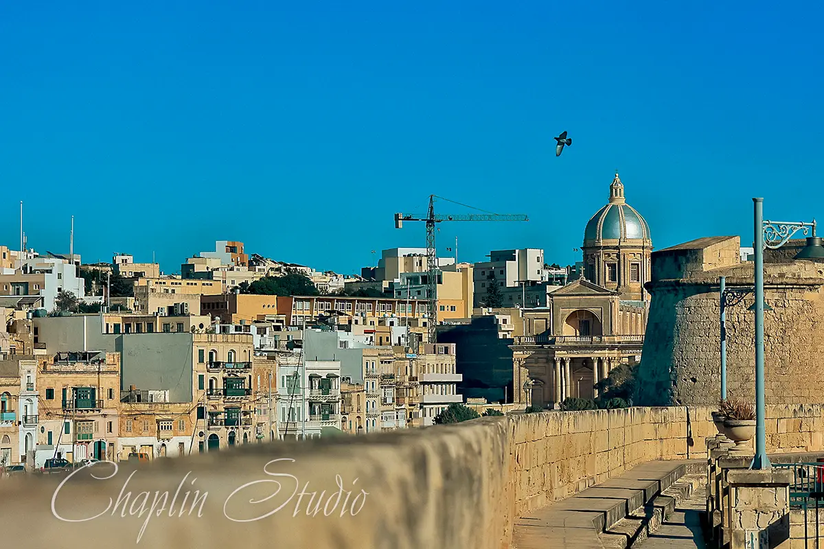Panoramic view of Valletta, Malta, showcasing a historic cityscape with a prominent dome, traditional buildings, stone fortifications, and a clear blue sky, captured by Chaplin Studio.