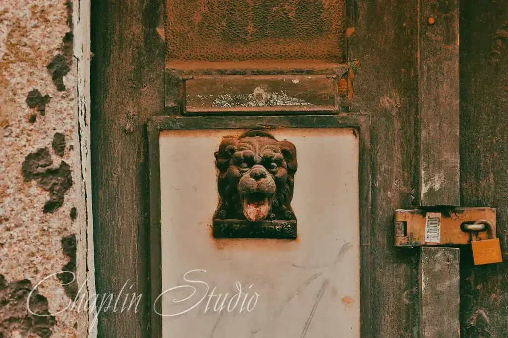 Close-up of a rustic, weathered wooden door with an ornate lion head door knocker, a mail slot, and a large, old padlock, showcasing vintage details captured by Chaplin Studio.
