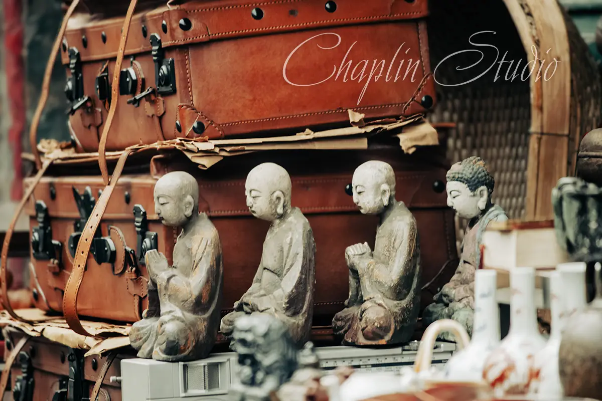 A collection of vintage brown leather suitcases stacked behind a row of small, meditating Buddha statues, with other antique items in a market setting, captured by Chaplin Studio.