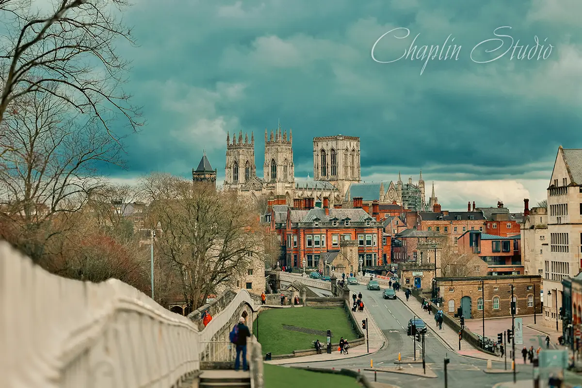 A panoramic view of York, England, featuring the majestic York Minster cathedral towering over historic city buildings, a bridge, and a street with people, all under a dramatic cloudy sky, captured by Chaplin Studio.