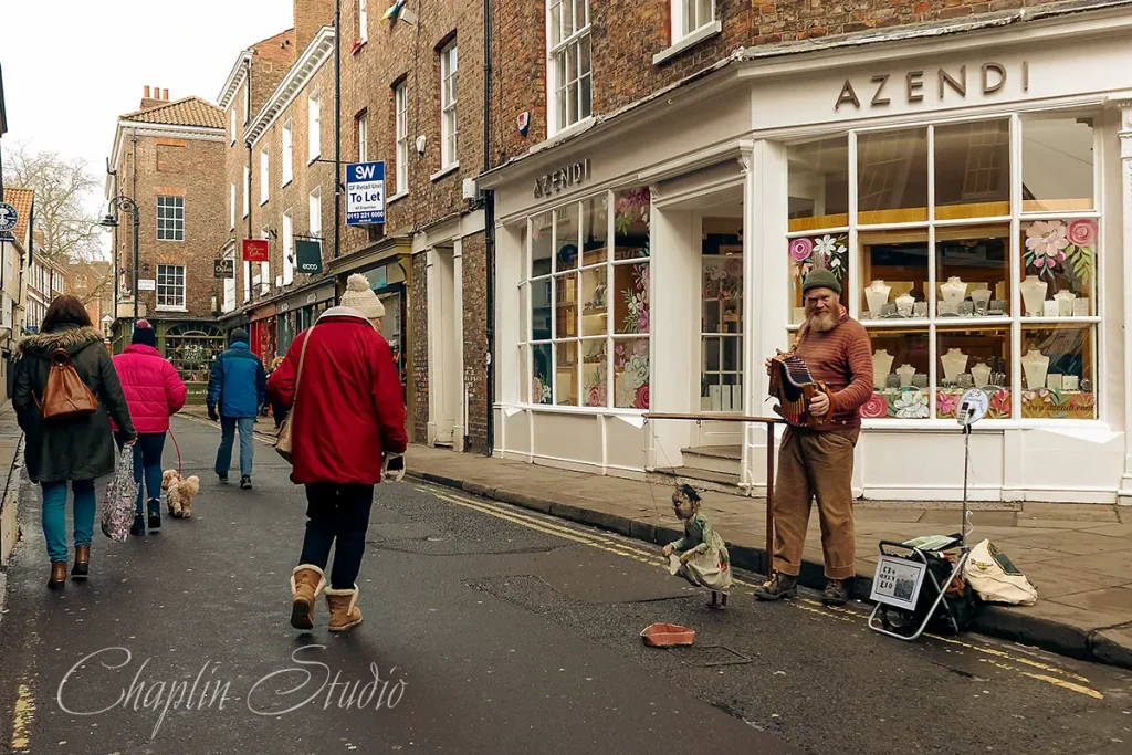 A lively street scene in York, England, featuring a street performer playing an accordion outside a jewellery store named 'AZENDI', with pedestrians and historic brick buildings, captured by Chaplin Studio.