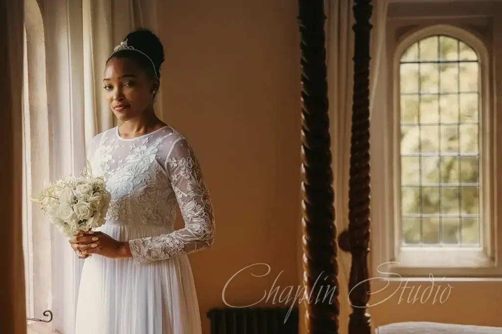 A beautiful bride in a white lace gown holds a bouquet by a window, captured in an elegant wedding portrait by Chaplin Studio in Yorkshire.