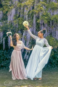 Two joyful bridesmaids in elegant dresses celebrating under blooming purple wisteria in a garden wedding scene