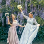 wo women in elegant dresses holding bouquets, celebrating joyfully in a lush garden setting with purple wisteria, ideal for wedding photography by Chaplin Studio in Yorkshire.