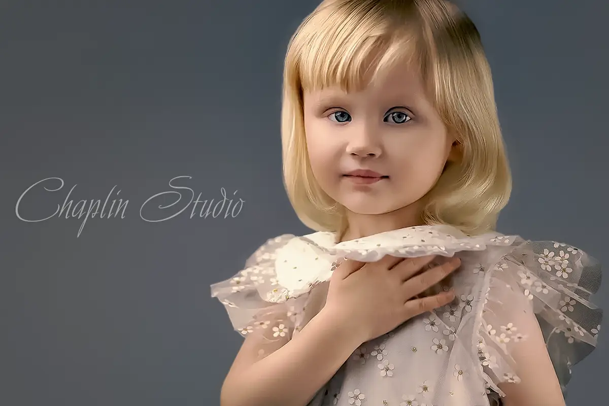 A young child with blonde hair and blue eyes, wearing a white dress, captured in a beautiful and innocent portrait by Chaplin Studio in Yorkshire.