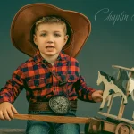 A young boy in a cowboy hat and plaid shirt, holding a wooden toy with horses, captured in a charming child portrait by Chaplin Studio in Yorkshire.