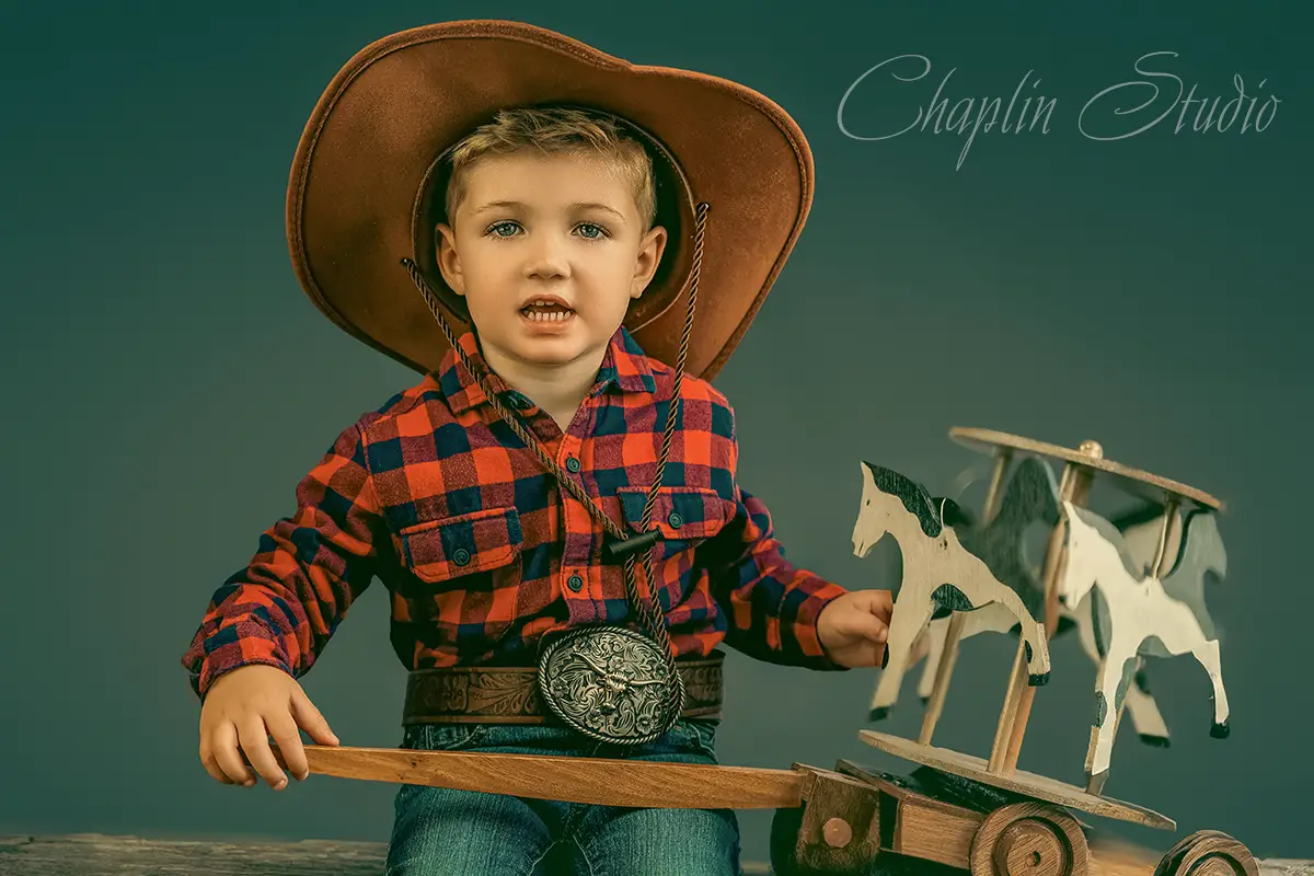 A young boy in a cowboy hat and plaid shirt, holding a wooden toy with horses, captured in a charming child portrait by Chaplin Studio in Yorkshire.