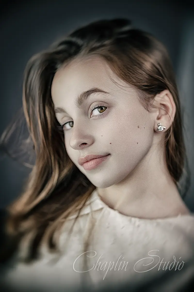 A close-up studio portrait of a young person with long hair, wearing a light-colored top, looking subtly at the camera, captured by Chaplin Studio in Yorkshire.
