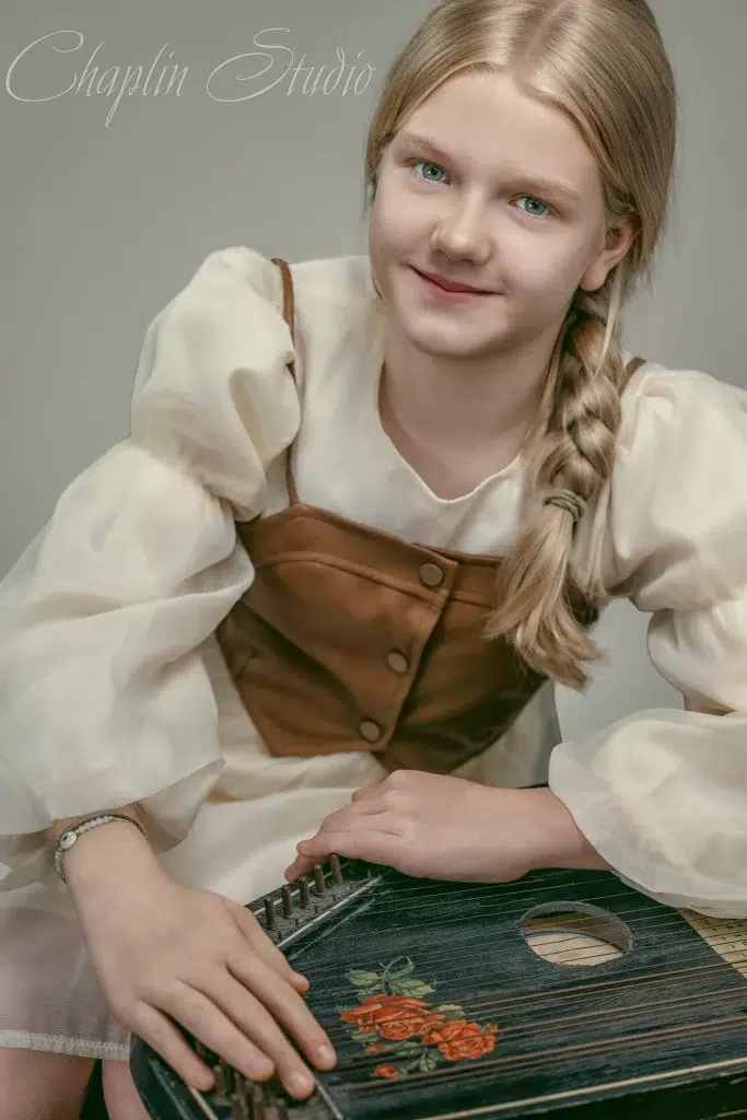 A young girl with blonde hair and a braid, wearing a historical-style dress and holding a stringed instrument, captured in a unique child portrait by Chaplin Studio in Yorkshire.