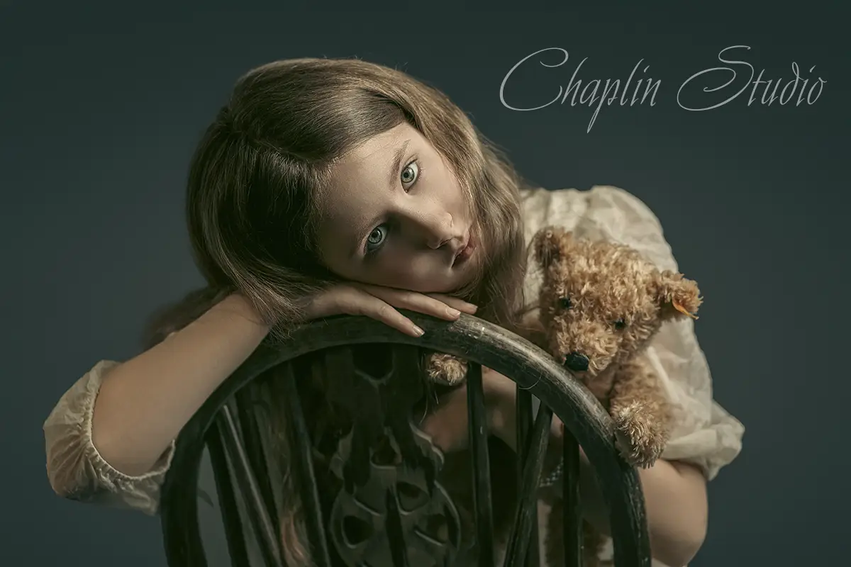 A young girl with a thoughtful expression, holding a teddy bear and resting on a wooden chair, captured in a heartfelt child portrait by Chaplin Studio in Yorkshire.