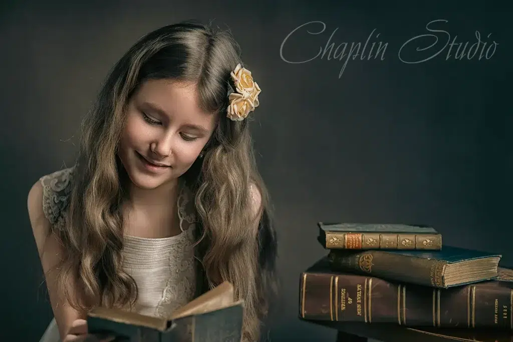 An artistic studio portrait of a child with long, wavy hair and a flower clip, happily reading a vintage book next to a stack of old books, captured by Chaplin Studio in Yorkshire.