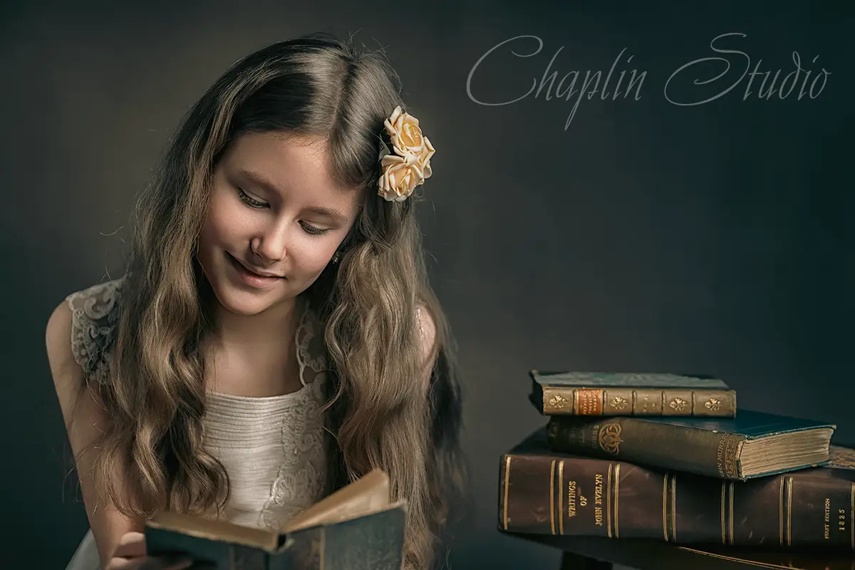 An artistic studio portrait of a child with long, wavy hair and a flower clip, happily reading a vintage book next to a stack of old books, captured by Chaplin Studio in Yorkshire.