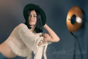 A creative studio portrait of a woman with dark hair wearing a stylish hat and a sheer top, leaning on a chair, captured by Chaplin Studio in Yorkshire.