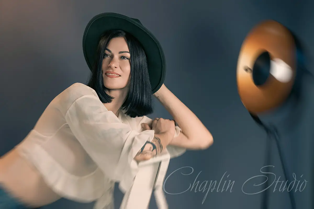 A creative studio portrait of a woman with dark hair wearing a stylish hat and a sheer top, leaning on a chair, captured by Chaplin Studio in Yorkshire.