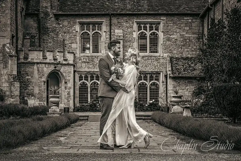 A couple embraces in front of a historic stone manor, captured in a timeless black and white wedding photograph by Chaplin Studio in Yorkshire.