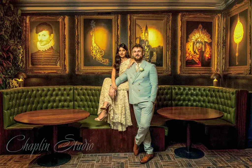 A stylish couple poses on a green leather booth in an ornate room with framed art, perfect for elegant wedding or portrait photography by Chaplin Studio in Yorkshire.