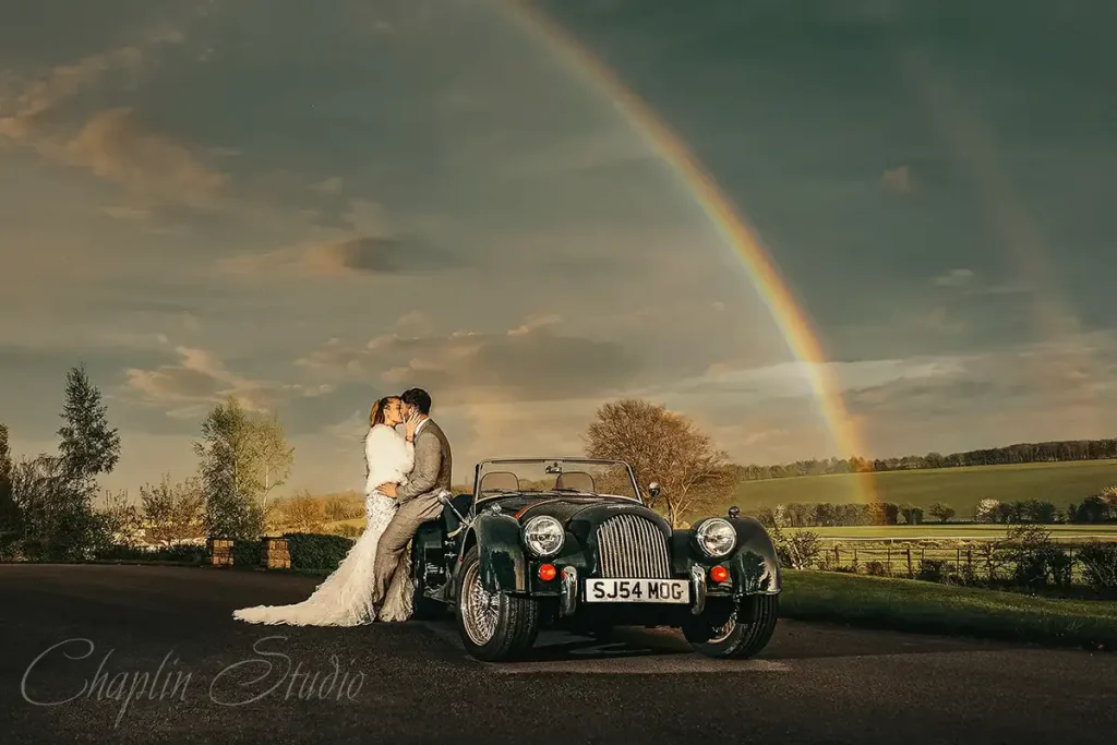 A newlywed couple shares a kiss by a vintage car under a vibrant rainbow in a picturesque Yorkshire landscape, captured by Chaplin Studio for wedding photography.