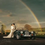 A newlywed couple shares a kiss by a vintage car under a vibrant rainbow in a picturesque Yorkshire landscape, captured by Chaplin Studio for wedding photography.