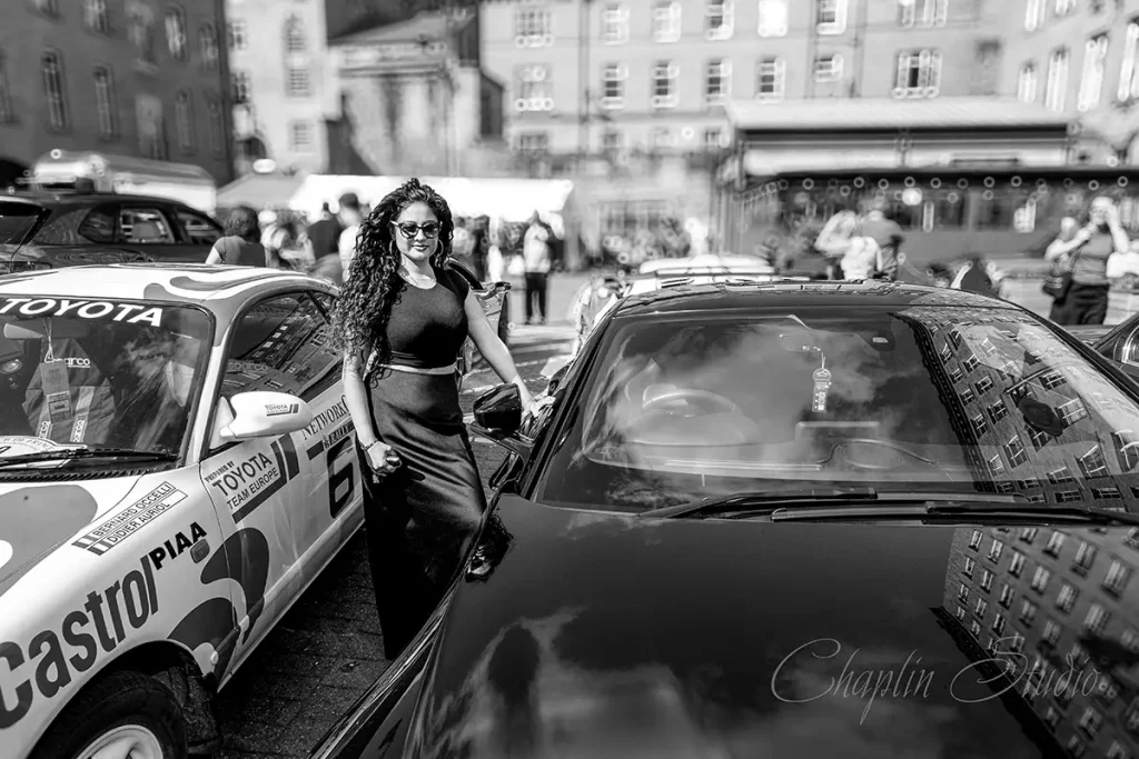 A woman in sunglasses and a dark outfit standing between two classic cars, one a Toyota rally car, at an outdoor event captured by Chaplin Studio.