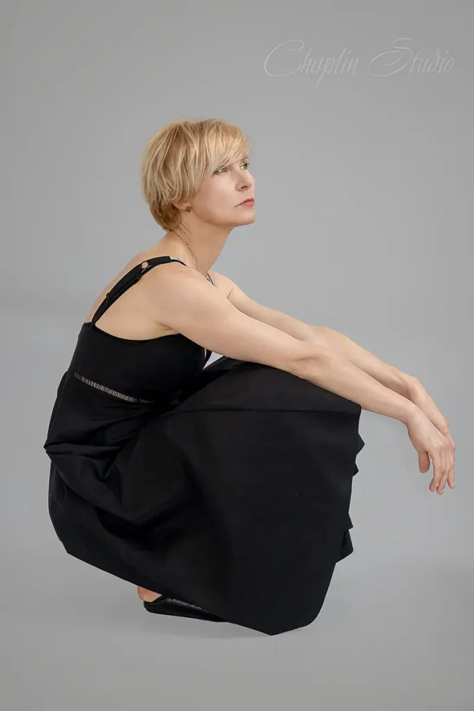 A creative studio portrait by Chaplin Studio of a woman with short blonde hair, wearing a black dress, squatting and looking thoughtfully to the side against a grey background.