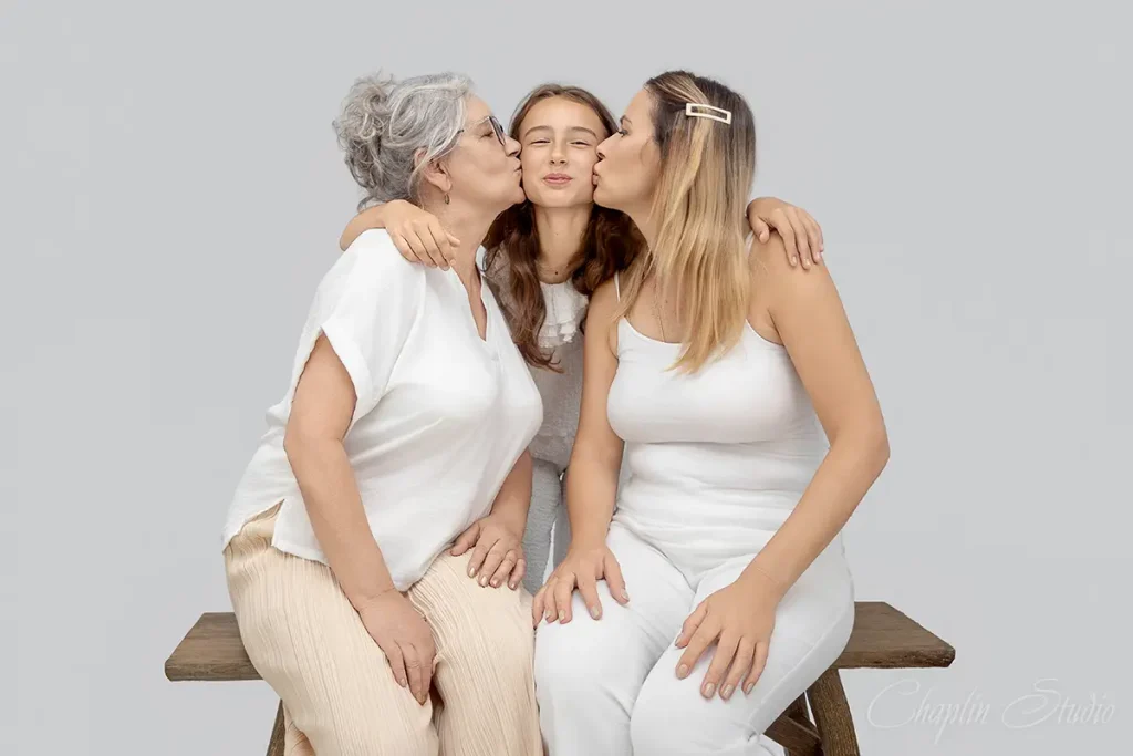 Three generations family portrait — grandmother, mother, and daughter sitting and sharing a kiss — natural light studio photo by Chaplin Studio