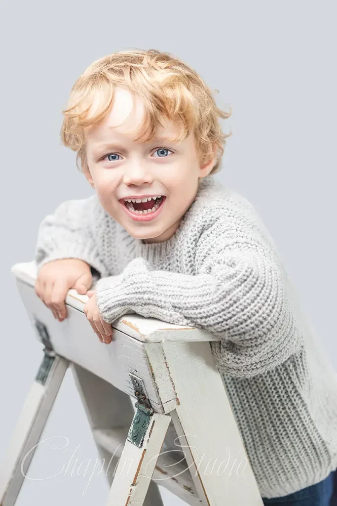 Portrait of a happy, smiling boy with curly hair sitting on a chair. Children's photography by Chaplin Studio in Halifax, UK.