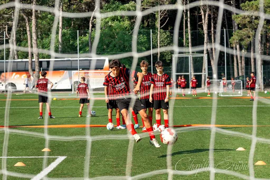 Youth football players in black and red striped kits training on a sunny pitch, viewed through a goal net, at a Milan Sporteventi camp.