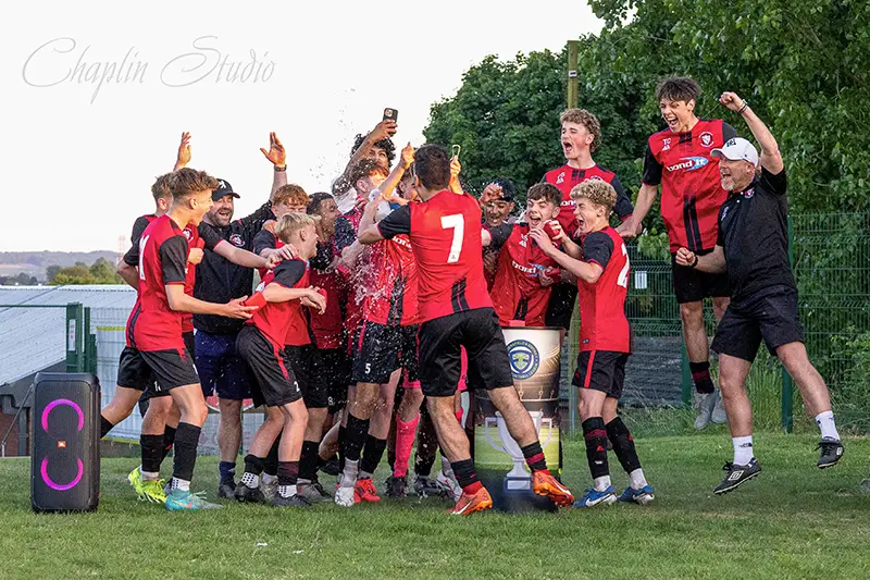 A youth football team in red and black kits celebrating a match victory on the pitch, with players and a coach cheering and spraying water in excitement.