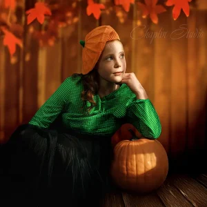 Red-haired girl in a pumpkin hat and green dress posing with a pumpkin in a Halloween-themed children’s portrait session at Chaplin Studio Yorkshire