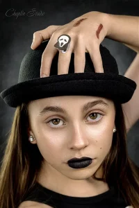 A close-up portrait of a person with brown hair, dark lipstick, and a black bowler hat, wearing a ghost ring on their finger. The photo has a spooky yet stylish Halloween vibe.